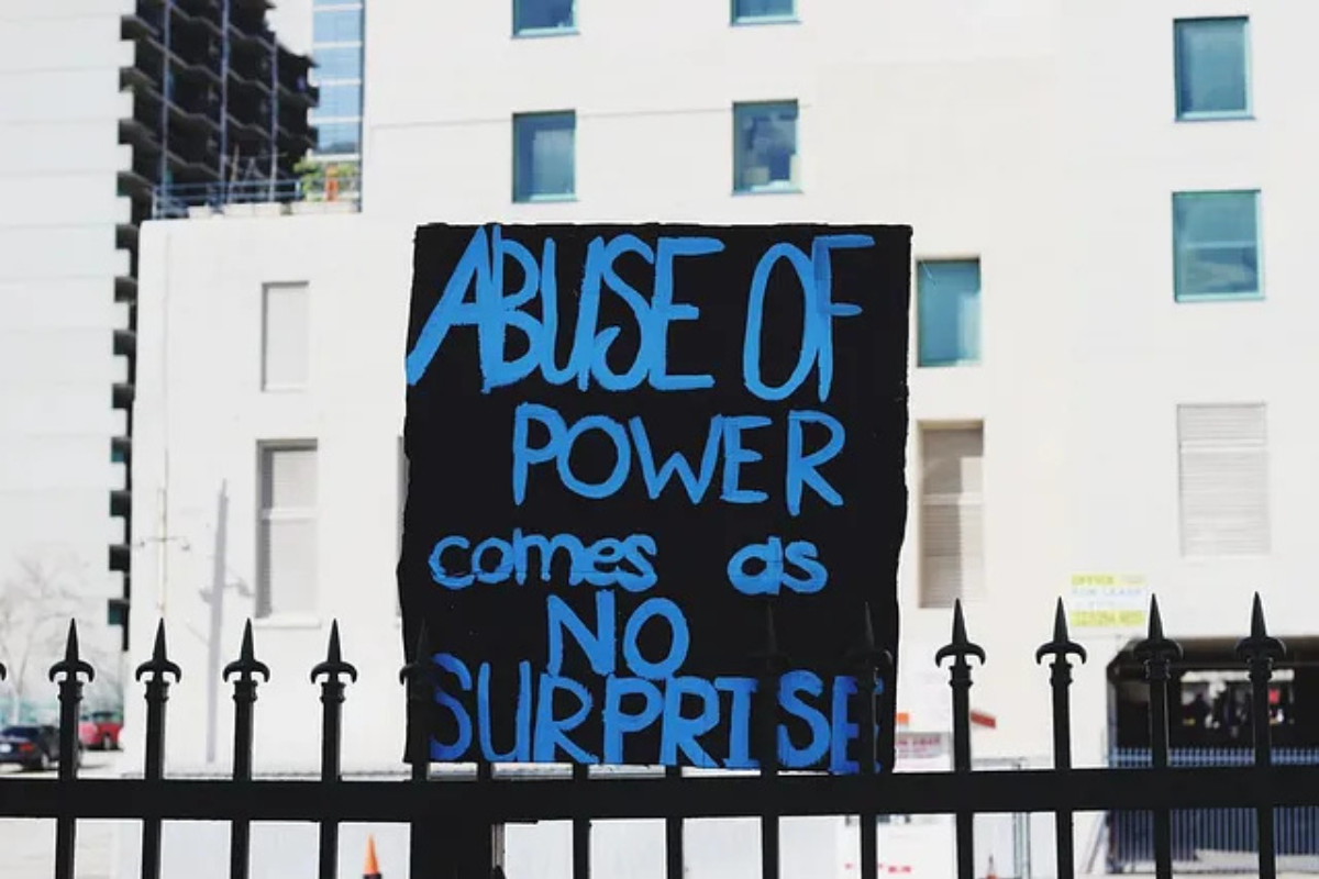 Sign reading 'Abuse of power comes as no surprise' in bold blue letters on a black background, mounted on a fence with buildings in the background.